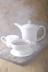 A white cup of hot tea and a white kettle on a wooden table.