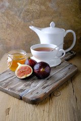 A white cup and a white teapot with tea, a jar of honey and fresh ripe figs on the cutting board on the table.