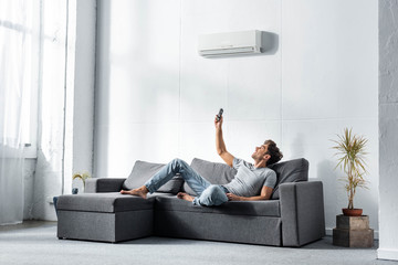 handsome man lying on sofa and switching on air conditioner - obrazy, fototapety, plakaty