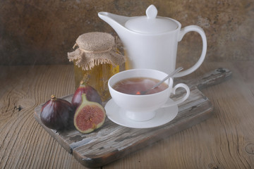 A white cup and a white teapot with tea, a jar of honey and fresh ripe figs on the cutting board on the table.