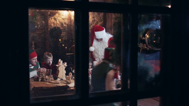 Santa Claus residence outside view through the window. Santa Claus and children with elves make out gifts