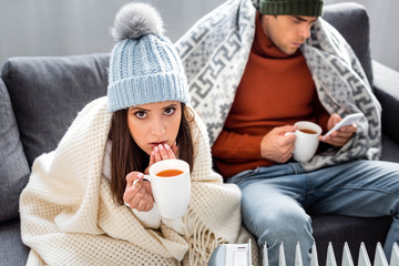 selective focus of attractive girlfriend with cup of tea warming up near heater