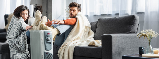 panoramic shot of attractive girlfriend and boyfriend covered with blankest warming up near heater