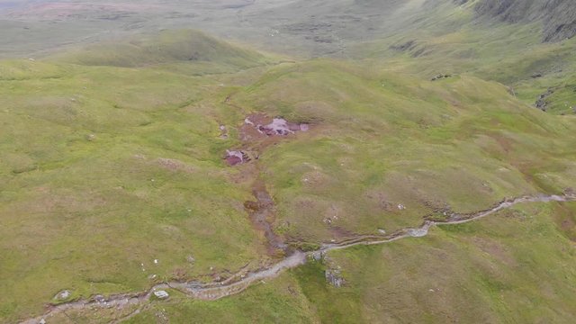 An aerial backward downward footage of a grassy mountain plateau rocky summit with small pond and mountain range in the background under a cloudy sky