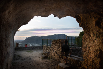 BAMBINO DI SPALLE CHE DA DENTRO UNA GROTTA NATURALE OSSERVA IL PANORAMA DEL MARE DI UNA CITTA' E DI UN MONTE