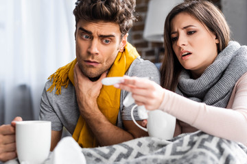 selective focus of sick girlfriend and boyfriend holding cups and looking at thermometer