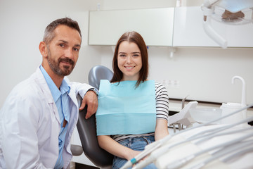 Obraz premium Attractive young woman sitting in a dental chair next to her dentist, copy space. Mature dentist and his female patient smiling to the camera after dental checkup