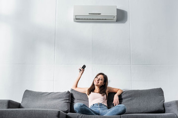 attractive and smiling woman switching on air conditioner with remote controller - obrazy, fototapety, plakaty