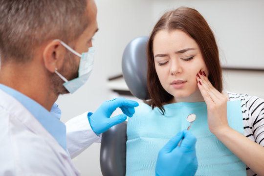 Young woman having toothache, sitting in a dental chair at the clinic. Female patient with terrible toothache visiting dentist