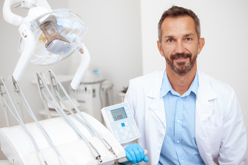 Handsome mature male dentist smiling cheerfully to the camera at his office. Dentistry, dental clinic, profession concept