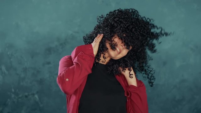 Young attractive woman with african curly hairstyle in pink shirt enjoying life and dancing at blue background. Hair flutters beautifully in slowmotion