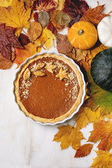 Traditional homemade autumn pumpkin pie for Thanksgiving or Halloween dinner served in ceramic dish with pumpkins and yellow autumn leaves over white marble background. Flat lay, space