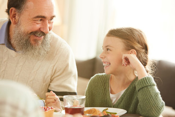 Happy little girl looking at her grandfather and smiling while sitting at the table together with him and have dinner