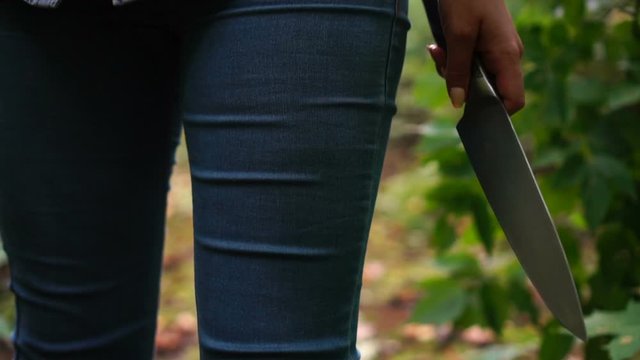 Woman with knife in her hand goes through green scary forest. Protective or criminal content.