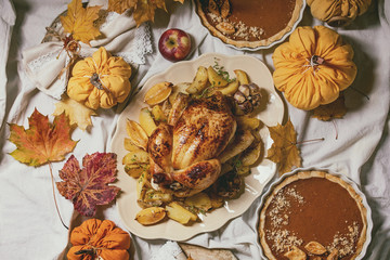 Thanksgiving or Halloween dinner with baked chicken with potatoes and lemons on big ceramic dish, pumpkin pies, plates, yellow autumn leaves, pumpkins as decorations over white table cloth. Flat lay