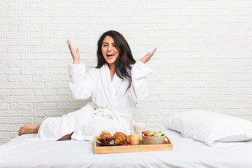 Obraz na płótnie Canvas Young curvy woman taking a breakfast on the bed receiving a pleasant surprise, excited and raising hands.