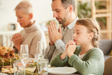 Family of three sitting at dining table with their eyes closed and praying before eating the meal at home