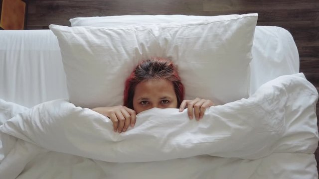 Asian girl with dyed hair appearing from under bed covers smiling and raising her eyebrows - Overhead view  of diverse millennial party girl waking up and being playful - bedroom and flirting concept