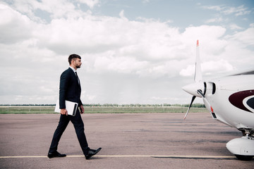 full length view of businessman in formal wear holding laptop and folder near plane in sunny day