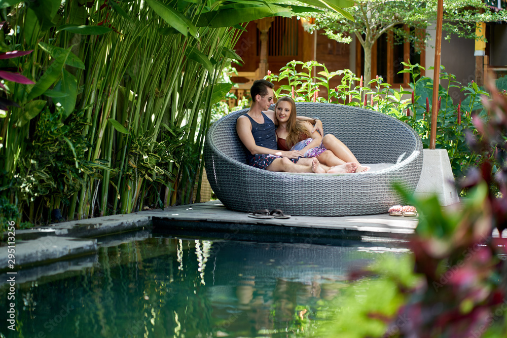 Wall mural candid shot of young caucasian couple lounging in modern chair near luxurious pool of tropical hotel