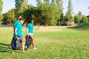 Team of eco activists leaving park after cleaning lawns. Young men and woman walking on grass, carrying rakes, plastic bags and talking. Garbage collection concept