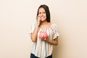 Young hispanic woman holding a piggy bank biting fingernails, nervous and very anxious.