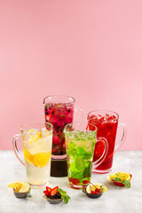 Group of lemonade jars with different ingredients and sorts of beverage at concrete textured table decorated with fruits and berries isolated at pink background.