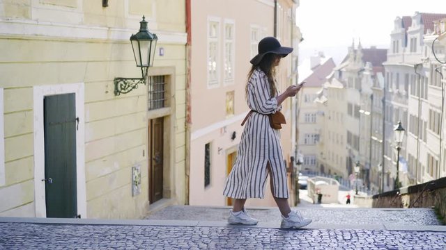 Trendy woman in hat walking at downtown of Prague.Lateral moving shot, side view girl in hat walking with smartphone on the stairway at blurred background residential buildings in downtown of Prague