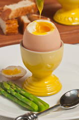 Top, front view, close up of a brown egg in a yellow porcelain egg holder with top cut off showing soft yolk with top on side and asparagus on a white round plate