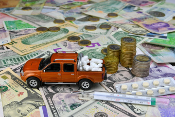 Thermometer and toy pickup with tablets in a cargo box on background of different countries currency banknotes. The concept of medical availability and increasing medical expenses.