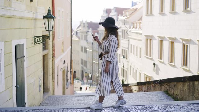 Trendy woman in hat walking at downtown of Prague. Side view young girl in hat taking selfie on smartphone, smiling and enjoying scenery while descending the stairway at old district Prague castle