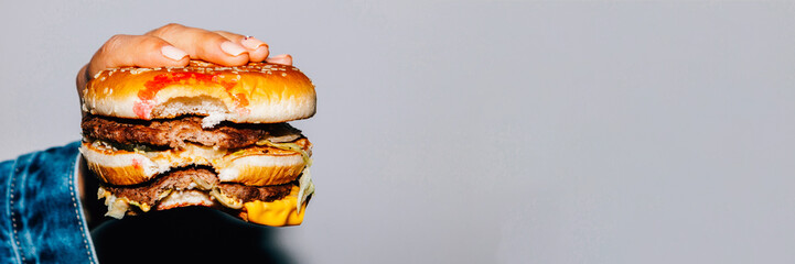 Unhealthy eating. Fast food. Bitten double cheeseburger in female hand over gray background.