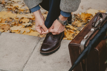 Gentleman is tying lace on brown shoe. Nearby is carpetbag