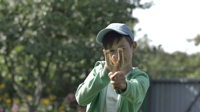 boy stands with a slingshot and aims at the camera outdors
