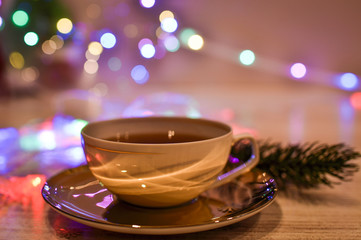 A cup of hot tea near the fireplace. Christmas garland with bright multi-colored lights near a white cup with coffee. Close-up photo. 