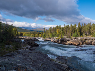 Beautiful northern landscape with wild glacier river Kamajokk, boulders and spruce tree forest in Kvikkjokk in Swedish Lapland. Summer sunny day, golden hour, dramatic clouds