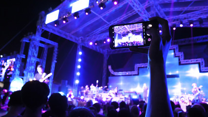 People live with mobile phone at a festival concert with crowd people raised hands and attending a...