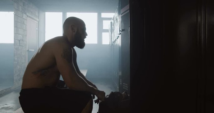 Young athletic shirtless Caucasian man sitting alone in dark gym locker room resting after workout, overcoming trouble.