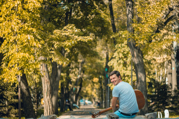 Fototapeta premium Young man sitting on the bench in the park playing on acoustic guitar with capo. Young attractive man enjoys live music in last sunny days autumn holiday. Retro lens used. Selective soft focus.