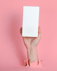 Female hand holds white box through torn pink paper. Minimalistic creative fashion concept