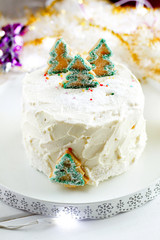 New Year's Christmas festive homemade cake with white butter cream glaze frost decorated with green Christmas tree cookies with garlands and lights on background. Bakery style cake for the celebration