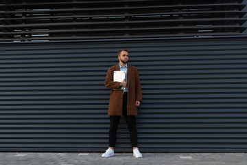 A young guy in a coat and with a laptop posing against a metal wall