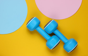 Plastic dumbbell on yellow background with blue pink pastel circles for copy space. Top view. Minimalistic sport concept. Top view