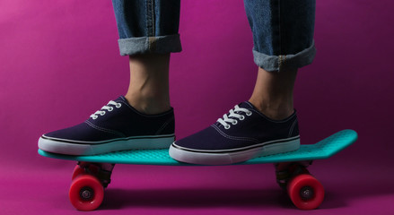 Youth hipster concept. Female legs in jeans and sneakers on cruiser board in the studio on purple...