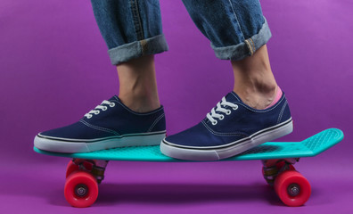 Youth hipster concept. Female legs in jeans and sneakers on cruiser board in the studio on purple...