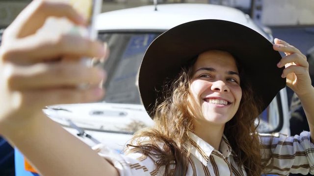 Charming girl walking around the city on a Sunny day. Close-up portrait elegant woman wearing hat and white dress posing on camera. Young lady taking selfie against blue hippie retro car on parking 