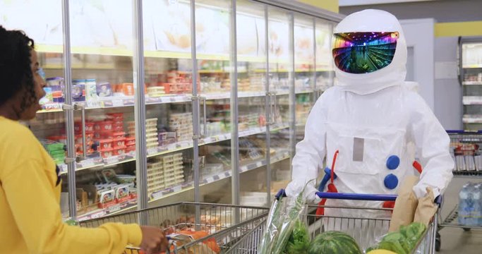 Funny spaceman going through the supermarket row with a trolley of food and waving his hand to the surprised young African American girl.