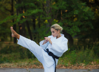 Attractive female master fighter in white kimono with black belt kicks outdoors. .