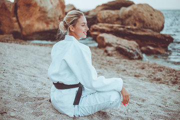 Young attractive karate woman in white kimono with black belt sitting on sand on wild beach with stones © splitov27