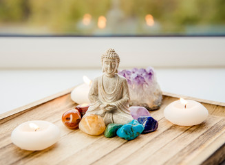 All seven chakra colors crystals stones around sitting Buddha figurine on natural wooden tray....